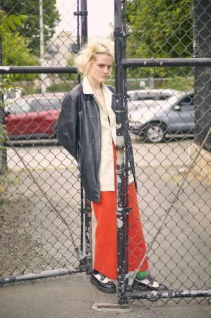 Blonde woman wearing a black leather jacket and red trousers leaning against a chain-link fence outdoors