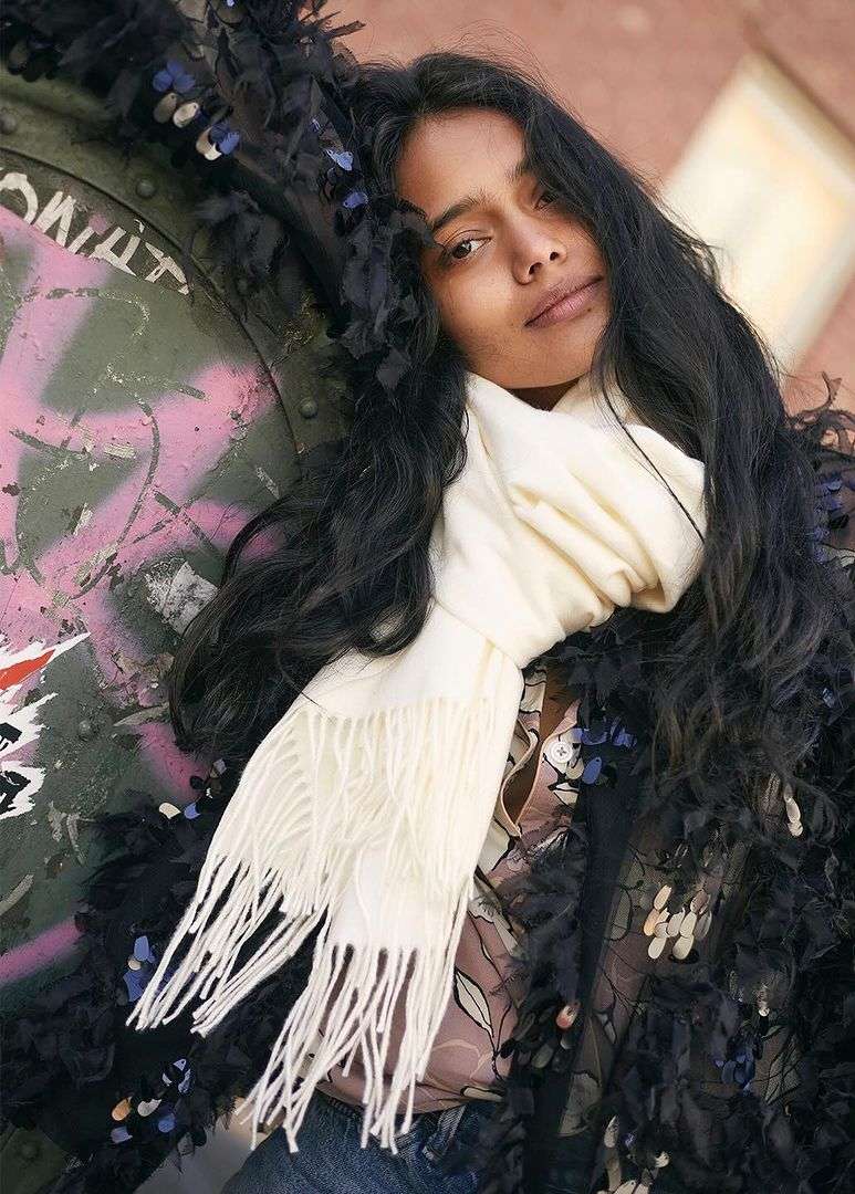Portrait of a woman with long dark wavy hair wearing a white fringed scarf outdoors against a graffiti wall