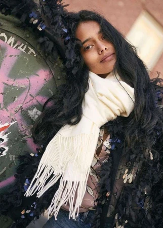 Portrait of a woman with long dark wavy hair wearing a white fringed scarf outdoors against a graffiti wall