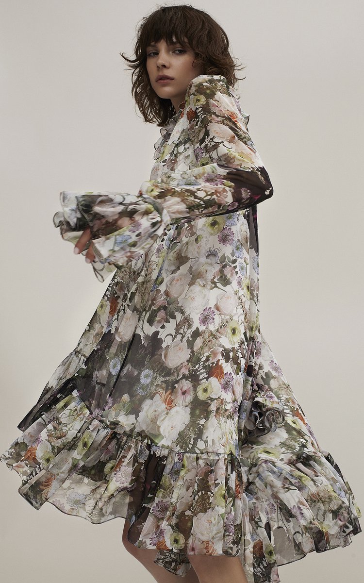 A woman poses indoors wearing a long floral dress with ruffles at the hem and cuffs, with a neutral background, demonstrating fashion.