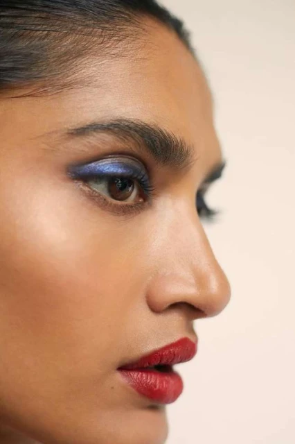 Close-up of an adult female's face in profile with blue eyeshadow and red lipstick against a neutral background in a beauty shot.