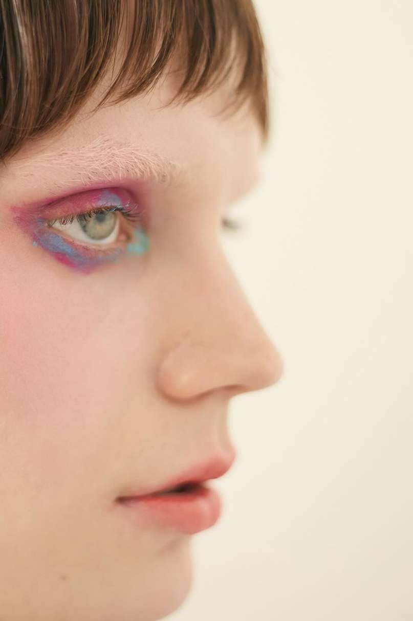 Profile view of a young adult woman with artistic makeup, featuring vibrant pink and blue eyeshadow, against a plain white background.