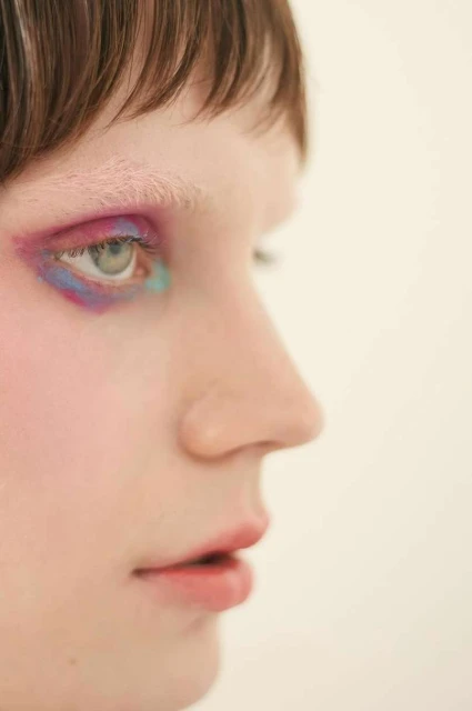 Profile view of a young adult woman with artistic makeup, featuring vibrant pink and blue eyeshadow, against a plain white background.