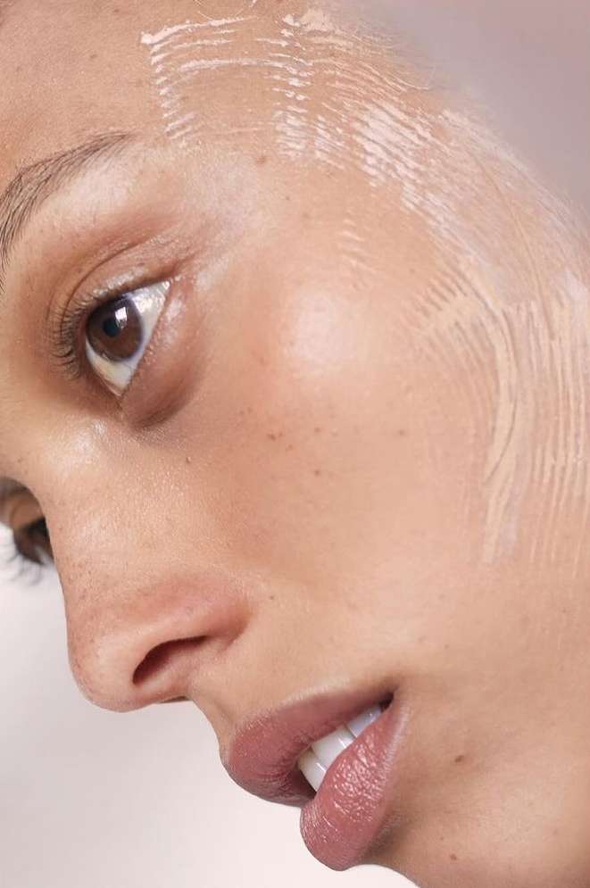 Close-up shot of a woman's face with clear skincare product applied, highlighting her skin texture, brown eye, and glossy lips.