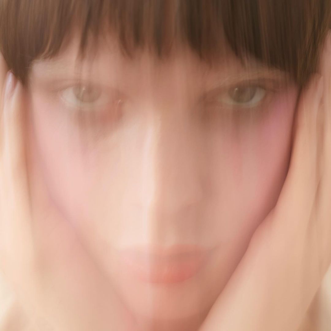 Abstract portrait of a young fair-skinned woman with dark hair, blurry face, holding hands to face, pink blush, and gazing at camera.
