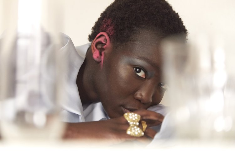 Close-up of a dark-skinned adult woman with pink-painted short hair, wearing a gold ring and light blue collared shirt against white.