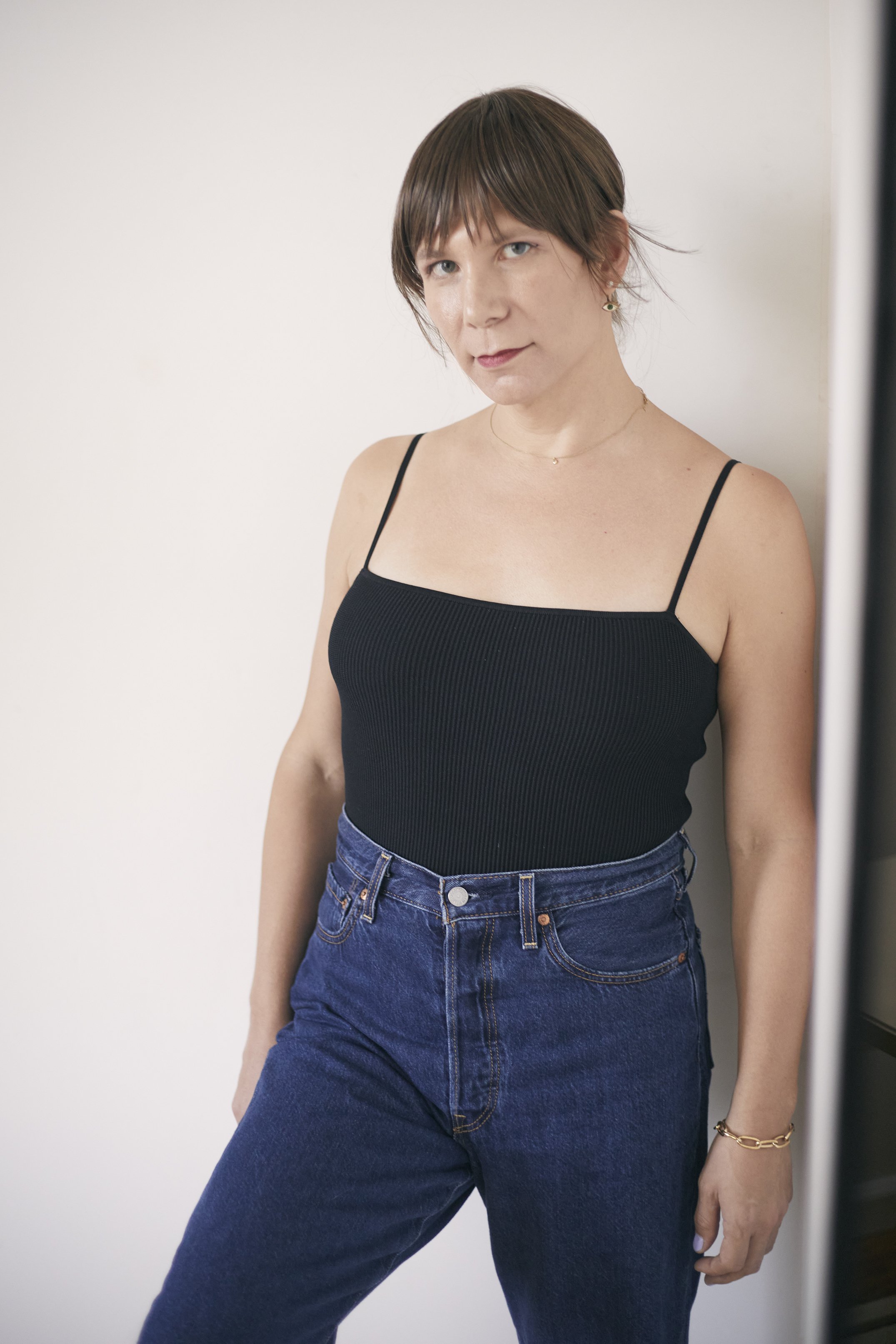 Woman with bangs wearing a black camisole and blue jeans posing indoors against a white wall.