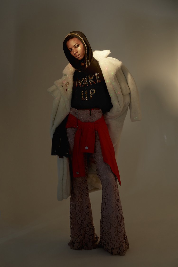 A fashionable young Black woman poses indoors wearing a hooded sweatshirt, red jacket, and lace pants with a white faux fur coat.
