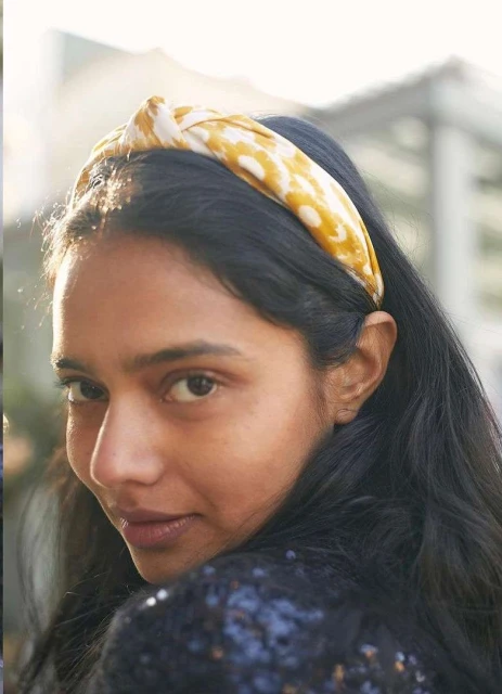 Portrait of an adult woman wearing a patterned yellow and white headband and a sparkly black top in an outdoor setting.