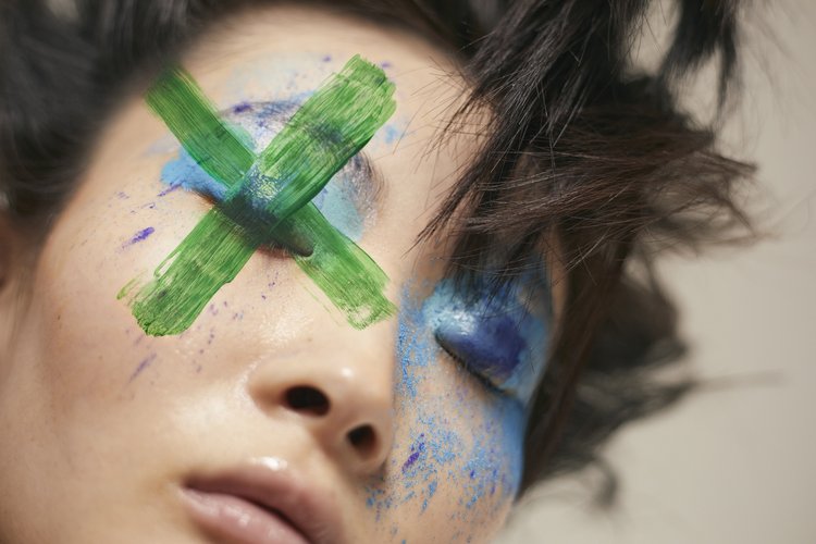 Close-up portrait of a woman with bold blue eyeshadow and a prominent green painted cross over her eyes by Viviane Black