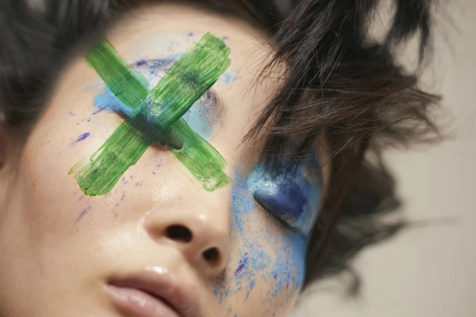 Close-up portrait of a woman with bold blue eyeshadow and a prominent green painted cross over her eyes by Viviane Black
