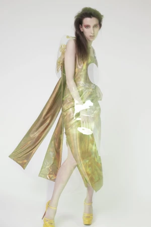 Model in shiny gold draped dress and yellow platform heels standing against a seamless white background.