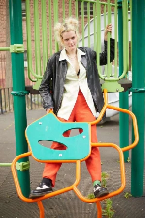 A young woman with blonde hair wearing a black leather jacket, blouse, and orange pants poses on a playground structure outdoors.