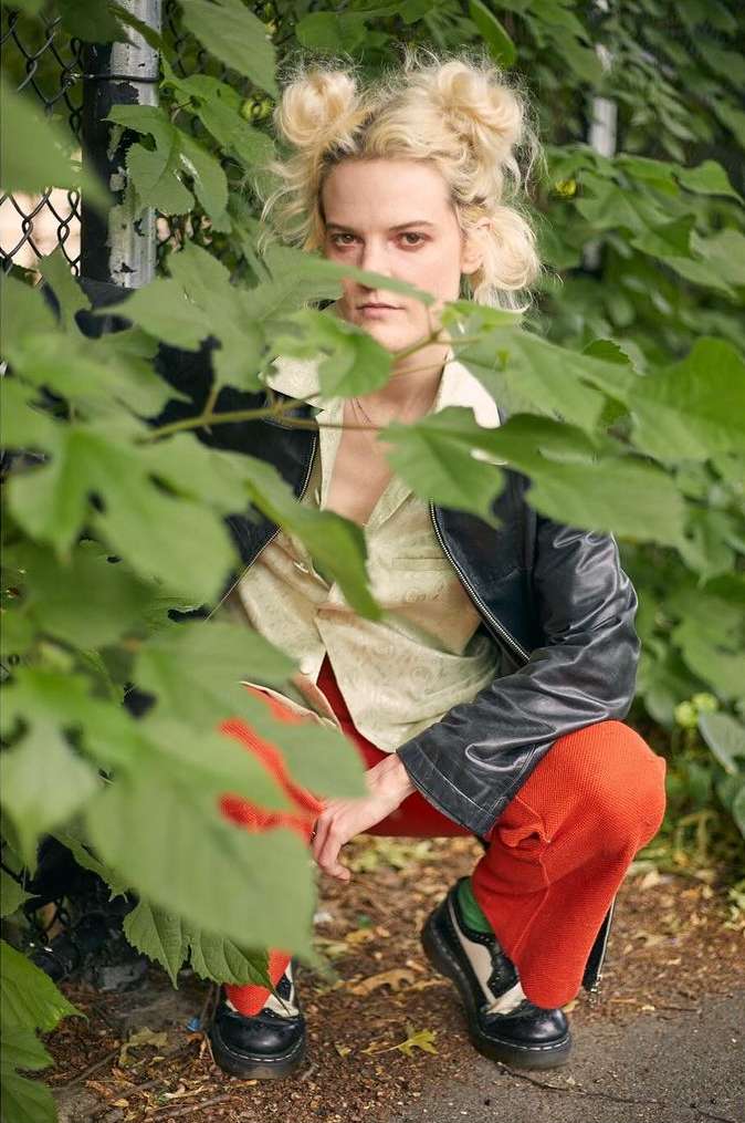 Blonde woman with space buns poses in a green outdoor setting, wearing an open black jacket, patterned blouse, and rust-colored pants.