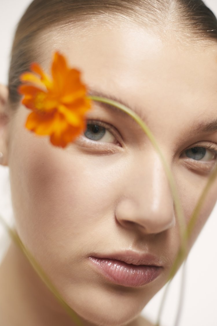 Close-up portrait of a woman with a bright orange flower held in the foreground across her face, photographed by Viviane Black.