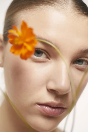 Close-up portrait of a woman with a bright orange flower held in the foreground across her face, photographed by Viviane Black.