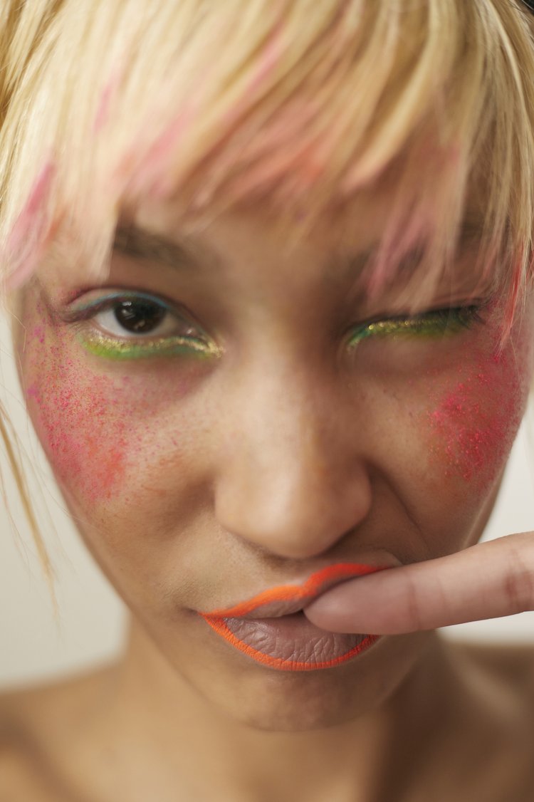 Close-up portrait of a young adult female winking with colorful makeup, orange outlined lips, blonde hair with pink highlights. Beauty and fashion.