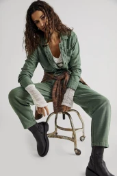 Woman in green jumpsuit seated on a metal stool wearing black boots and a plaid shirt tied at her waist against a white background.