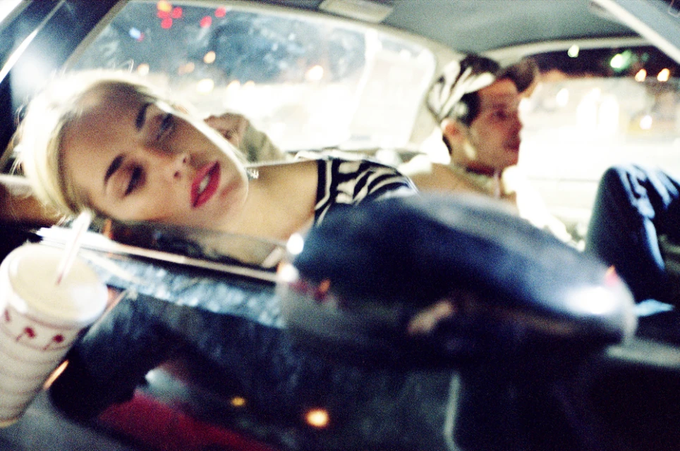 Woman resting in car with drink at night, with another person in the background; soft lighting and shallow depth of field.
