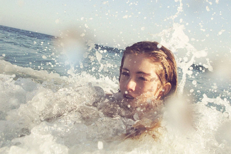 Woman in the ocean surrounded by splashing water, with pale complexion and light brown hair, captured with a shallow depth of field.