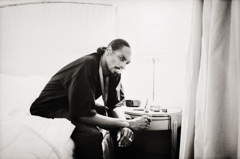 Black and white photo of a man sitting on a bed in a robe, next to a nightstand with an alarm clock and a lamp.