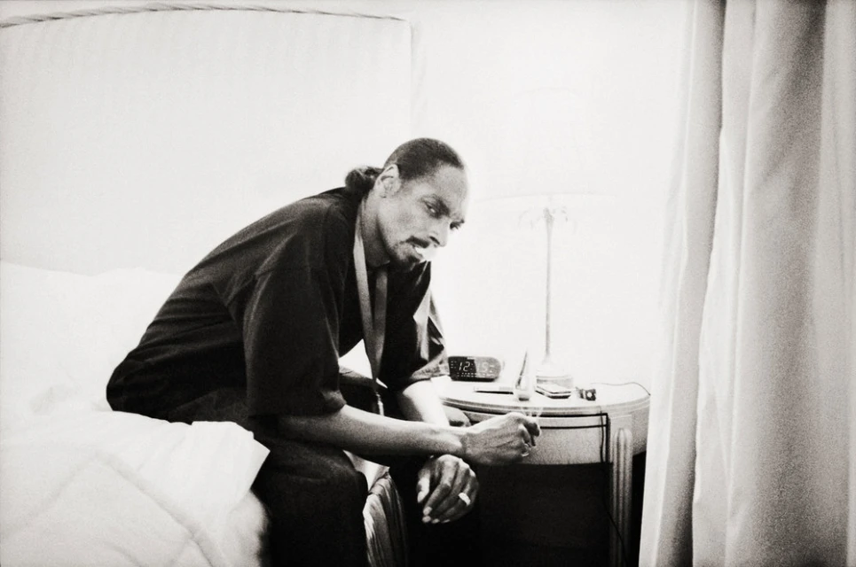 Black and white photo of a man sitting on a bed in a robe, next to a nightstand with an alarm clock and a lamp.