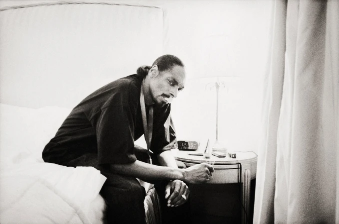 Black and white photo of a man sitting on a bed in a robe, next to a nightstand with an alarm clock and a lamp.