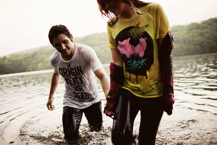 A young couple, soaked and smiling, walks through a lake, wearing graphic T-shirts, with trees and water in the background on an overcast day.