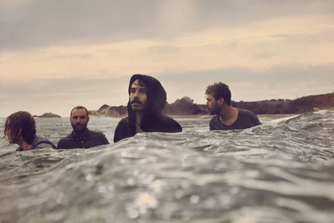 Four men standing in shallow ocean water with waves cresting in the foreground under an overcast sky.