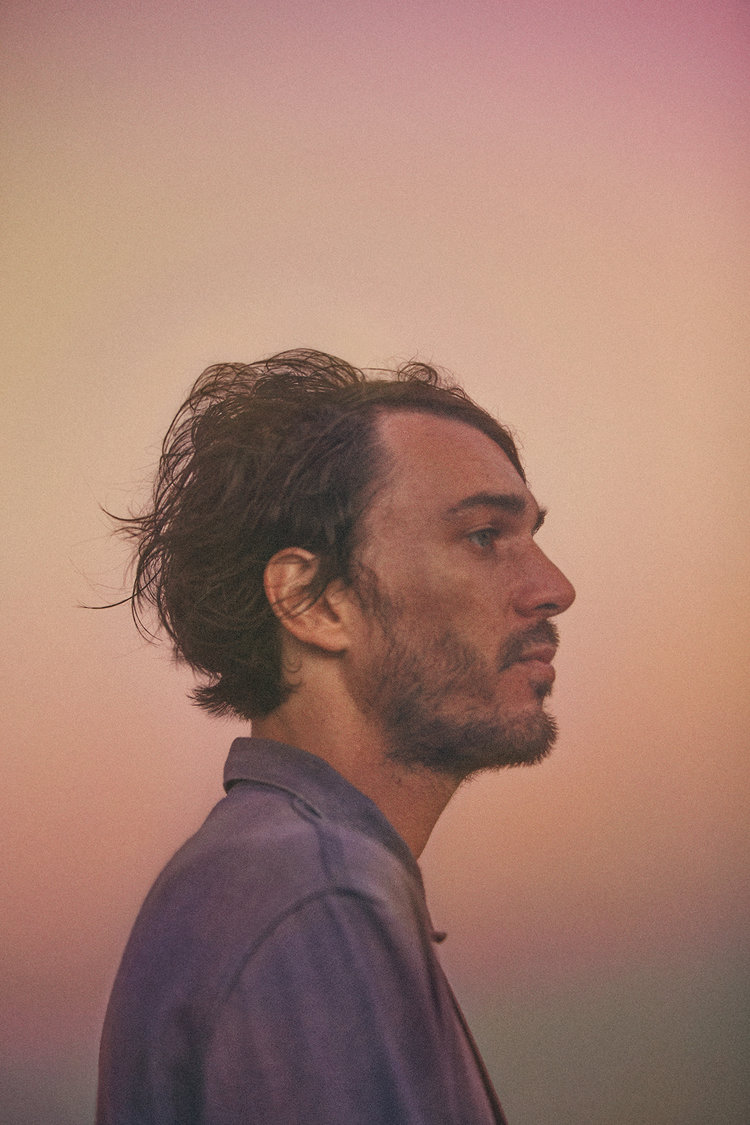Profile view of a man with dark hair and a beard, wearing a blue collared shirt against a gradient peach, pink, and yellow background.
