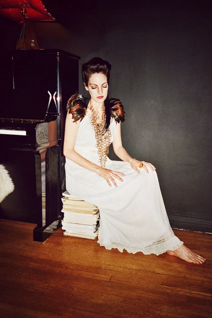 Woman in white gown poses indoors, seated on a stack of books next to a black piano with dark gray walls and a hardwood floor.