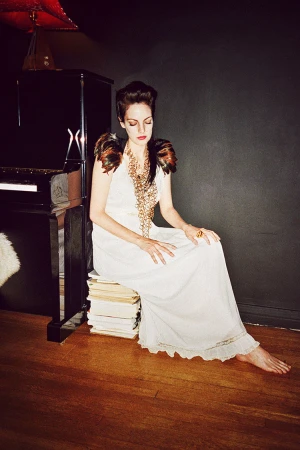 Woman in white gown poses indoors, seated on a stack of books next to a black piano with dark gray walls and a hardwood floor.