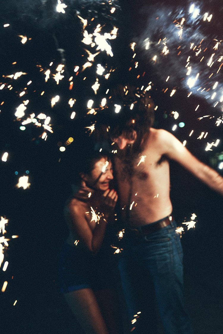 A shirtless man embraces a woman while sparklers create bright bursts of light in the dark, creating a romantic and festive atmosphere.