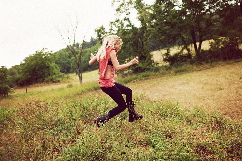 Blonde woman in pink sleeveless top and black jeans jumping through a grassy field by photographer Bryan Sheffield
