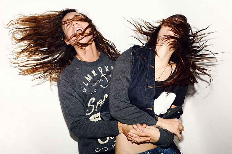 Two young adult women embrace with their hair blowing across their faces in a close shot against a white background in a studio.