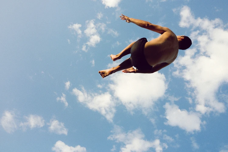 Adult male in mid-air, appearing to dive, against a vibrant blue sky dotted with fluffy white clouds on a sunny day.