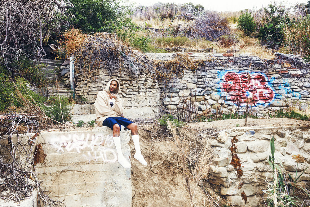 Adult male in athletic wear sitting on stone steps in a rural outdoor environment with vegetation and a stone wall behind him.