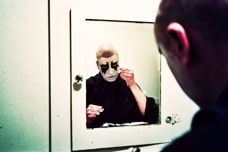 Reflection of bald adult male in bathroom mirror, applying black and white face paint in an indoor, domestic setting.