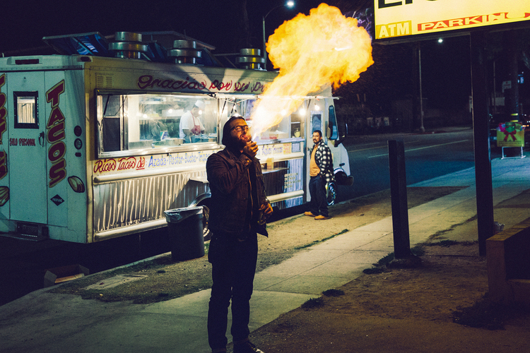 Man breaths fire in front of food truck at night. A second man stands nearby, as the flames reach high in the urban setting.