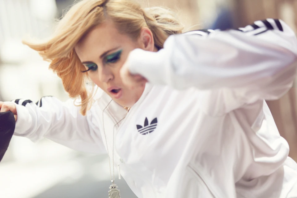 An adult female with blonde hair wearing a white Adidas track jacket and long pendant necklace, with vibrant green eyeshadow and pink lips.