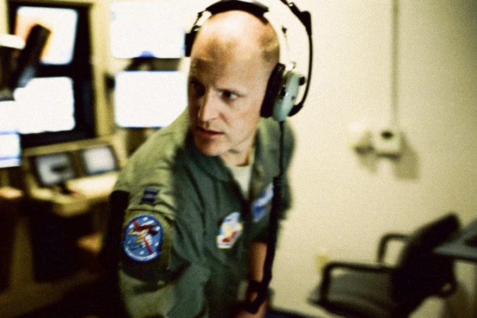 An adult male wearing a headset and green military uniform looks to the left in an indoor setting with monitors and control panels.