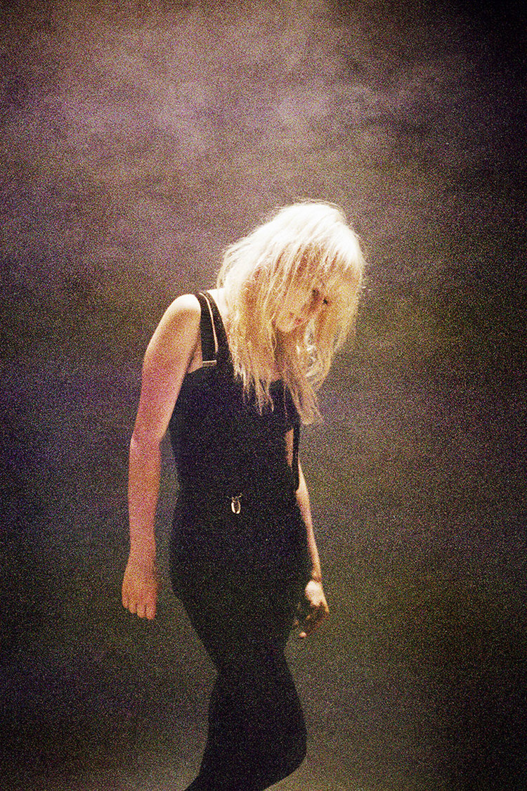 An adult female stands with her head tilted, illuminated by a stage light. She has blonde hair and wears a dark outfit.