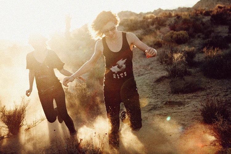 Two women run hand-in-hand across a sandy desert landscape at sunset, kicking up dust as they go.