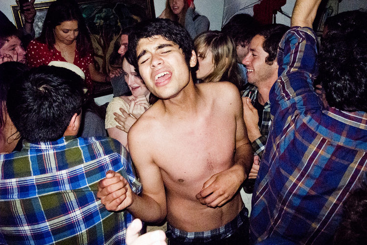 Indoor candid shot of a young adult singing at a party, surrounded by a crowd of people in a dimly lit room.