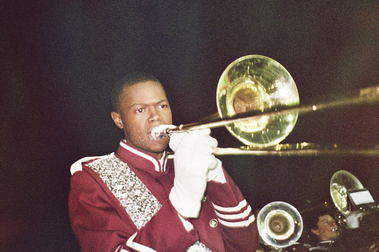 Musician plays trombone wearing marching band uniform against a dark backdrop with brass instruments visible in the background.