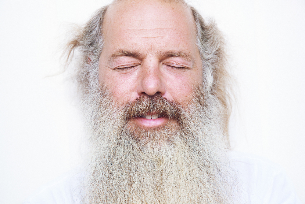 Close-up of an adult male with a long, white beard and closed eyes against a stark white background, focusing on his facial features.