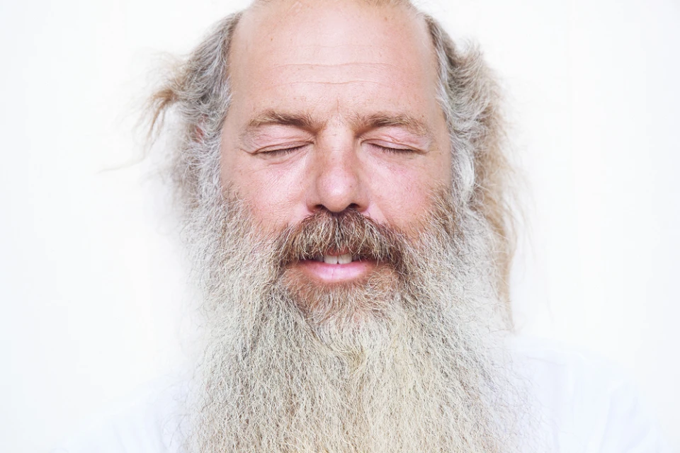 Close-up of an adult male with a long, white beard and closed eyes against a stark white background, focusing on his facial features.