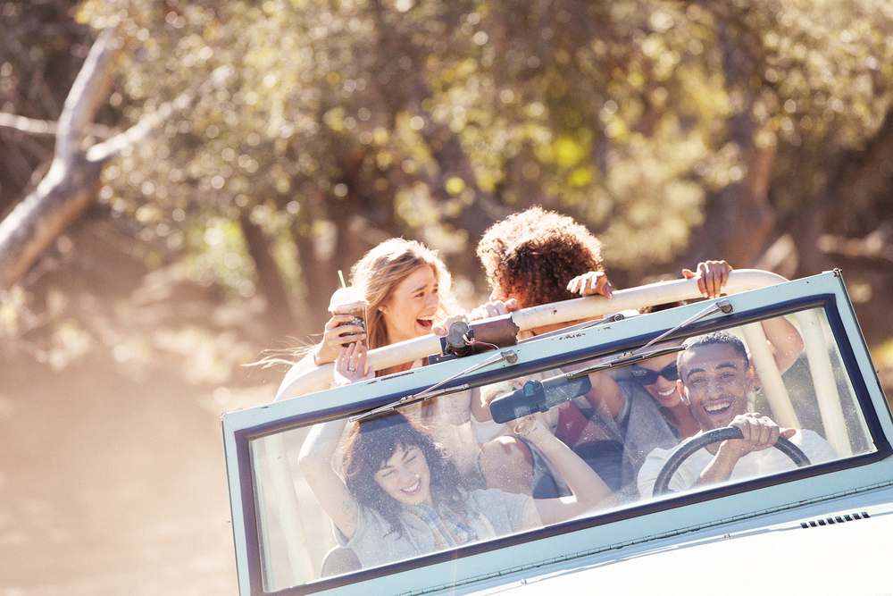 Four people ride in a jeep on a sunny day. The open-air vehicle moves along a tree-lined path with happy passengers. 