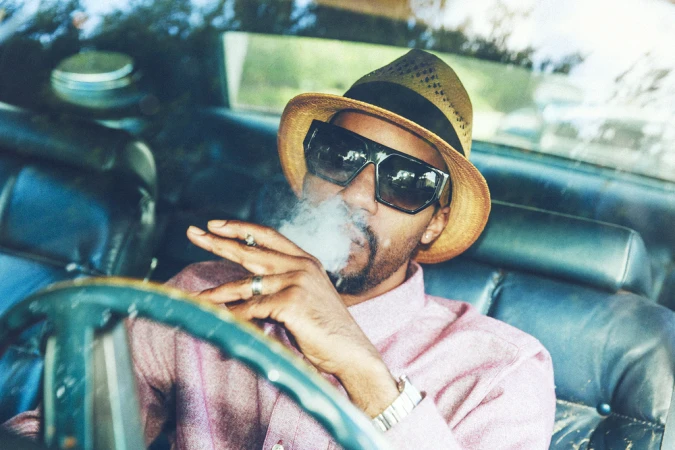 Man wearing sunglasses and a fedora hat smoking a cigarette inside a car, photographed by Bryan Sheffield.