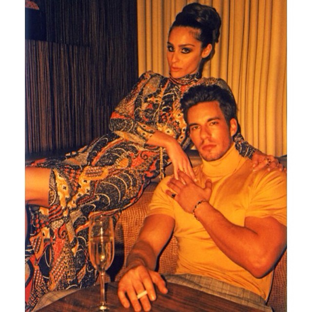 Stylish portrait of a woman in an ethnic dress and a man in a yellow turtleneck sitting on a sofa in an indoor setting.